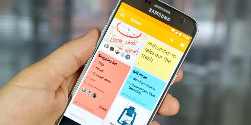 A hand holding a Samsung smartphone with the Google Keep app open, showcasing multiple interactive notes: a handwritten-style reminder (“Gotta check the attic!”), a shopping list, gift ideas, and a to-do item (“remember to take out the trash”). The interface demonstrates real-world use for personal organization and collaboration