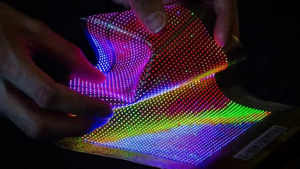 Flexible LED textile panel being bent by hands, displaying multicolored illuminated pixels to demonstrate electronic fabric flexibility.
