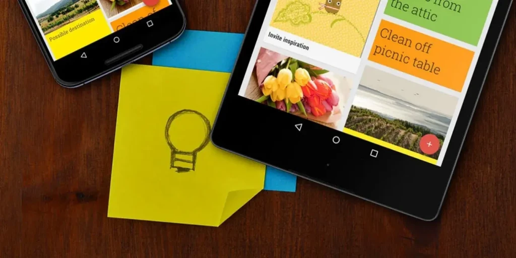 A tablet and smartphone side by side on a wooden surface, both showing Google Keep’s colorful, image-rich notes, including “Invite inspiration” with tulips and “Clean off picnic table.” A physical yellow sticky note with a hand-drawn lightbulb sits between them, blending digital organization with tactile creativity