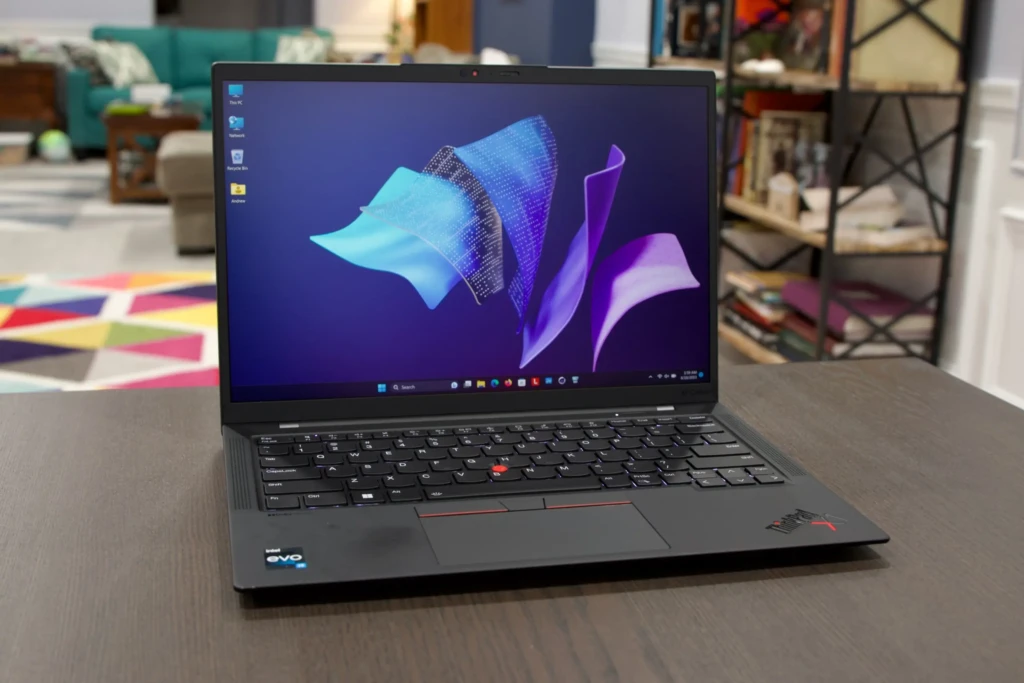 A Lenovo ThinkPad X1 Carbon laptop open on a dark wooden table in a living room setting, displaying the Windows 11 default wallpaper. The screen shows desktop icons and taskbar; an Intel Evo badge is visible on the palm rest