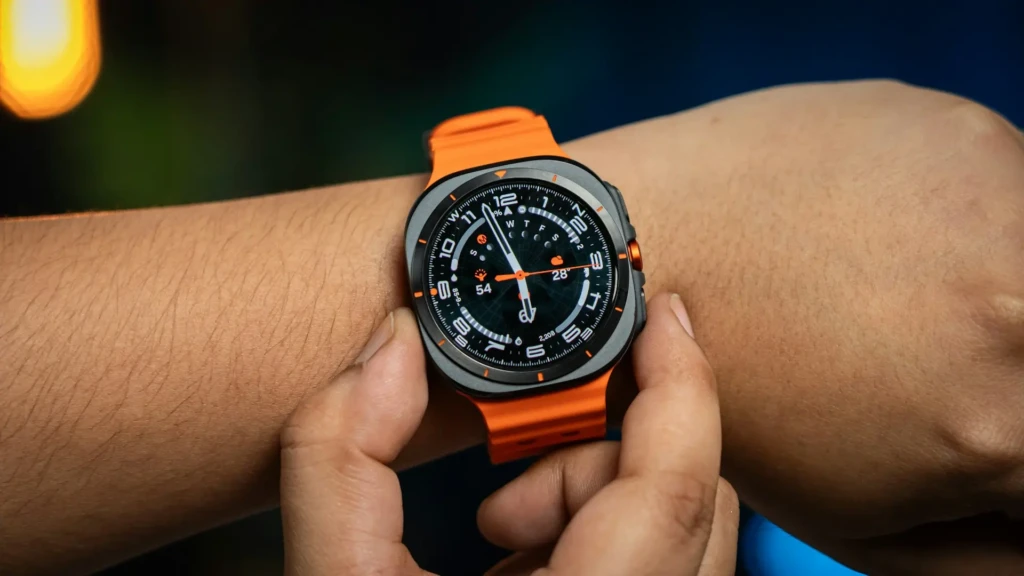 A close-up of a person adjusting the Samsung Galaxy Watch Ultra with an orange strap on their wrist. The watch face shows time, heart rate, weather, and step count, under warm ambient lighting, suggesting evening or outdoor use