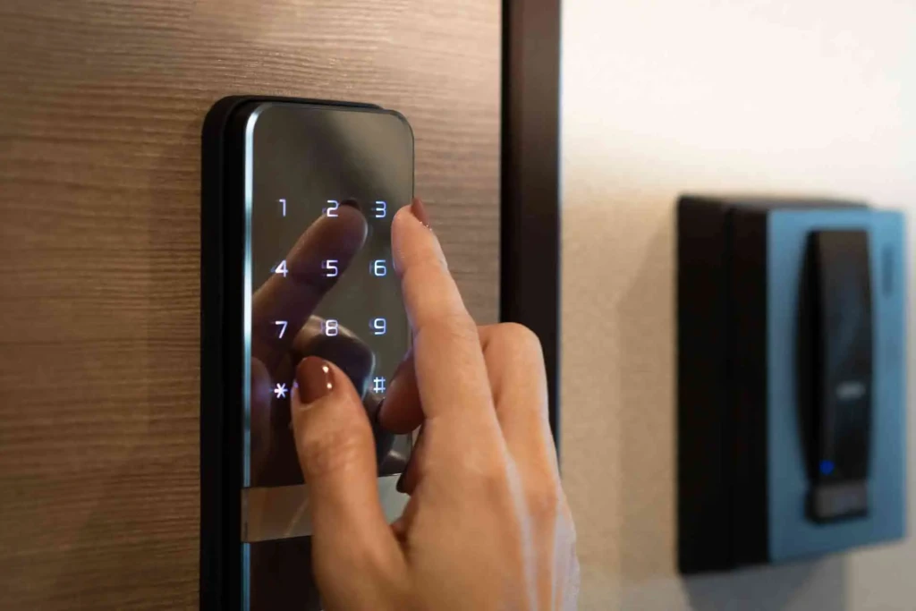 A close-up of a hand with brown nail polish entering a code on a sleek black digital keypad smart lock mounted on a wooden door, with a card reader visible in the background, illustrating modern keyless entry and security convenience.