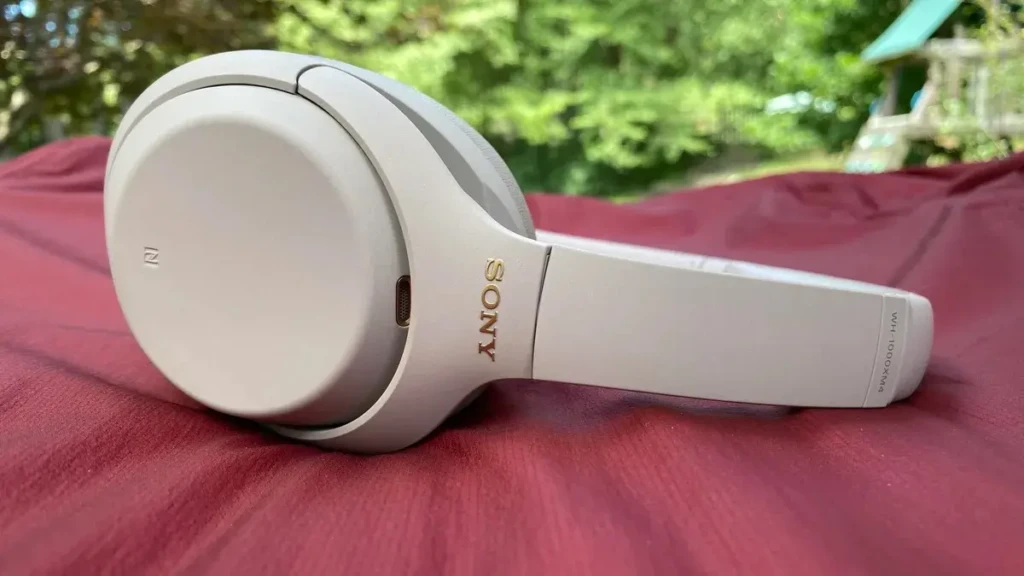 A side view of the cream-colored Sony WH-1000XM4 headphones resting on a red fabric surface outdoors, with green foliage softly blurred in the background, showcasing its elegant profile, USB-C port, and comfortable earcup design in a natural, relaxed setting.