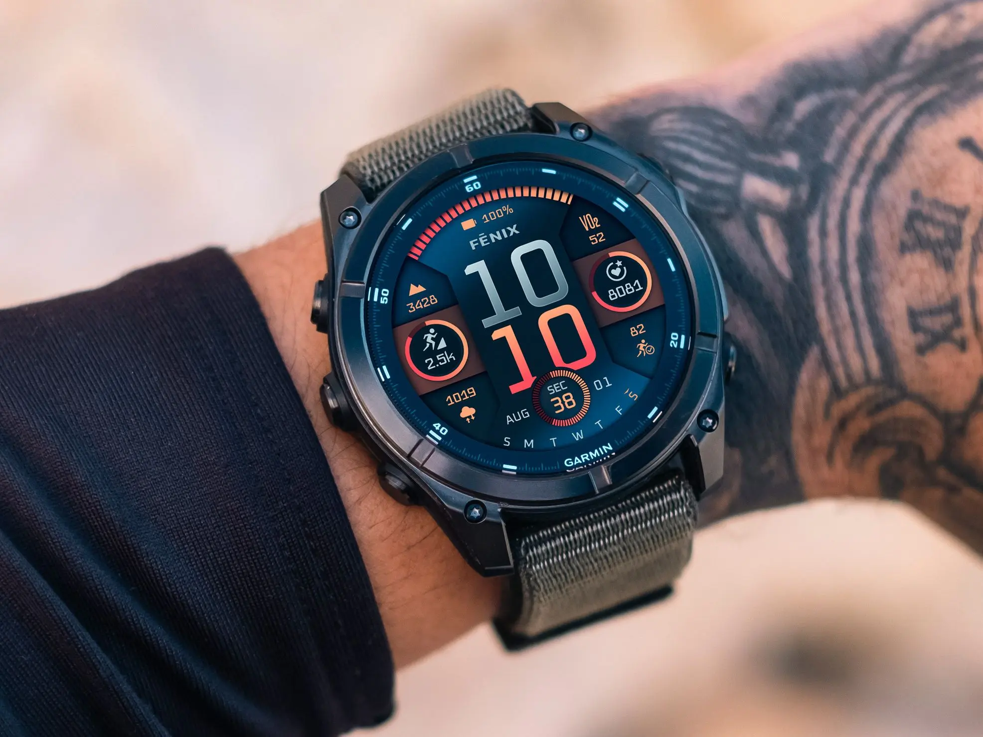 A close-up of a Garmin Fenix 8 on a person’s tattooed wrist, featuring a dark gray fabric strap. The vibrant digital watch face shows time (10:10), battery at 100%, VO₂ max (52), steps (8,081), and active distance (2.5k), highlighting its detailed fitness tracking and bold, customizable display