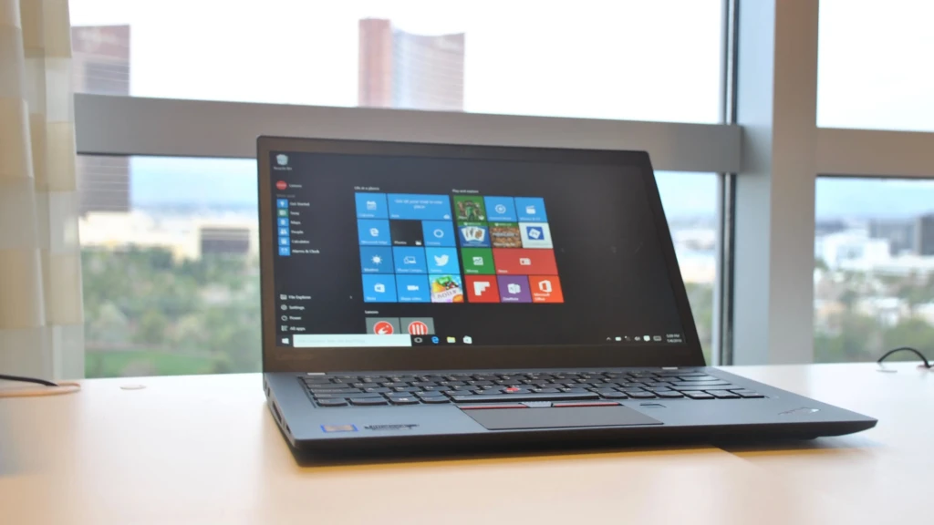 A Lenovo ThinkPad laptop sitting on a white desk near a large window with a cityscape view, displaying the Windows Start menu with colorful app tiles. The keyboard features the iconic red TrackPoint and trackpad