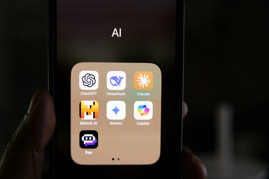 A hand holding a smartphone displaying a folder labeled “AI,” containing app icons for ChatGPT, DeepSeek, Claude, Gemini, Copilot, Mistral AI, and Poe, illustrating how DeepSeek fits into a broader ecosystem of popular AI tools on mobile devices.