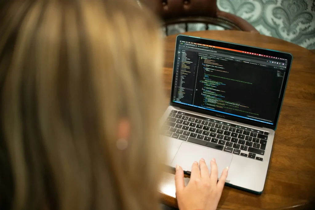 An over-the-shoulder view of a person with blonde hair typing on a MacBook Pro, which displays lines of JavaScript or TypeScript code in a dark editor, emphasizing hands-on coding and personal focus in a cozy, home-office setting.
