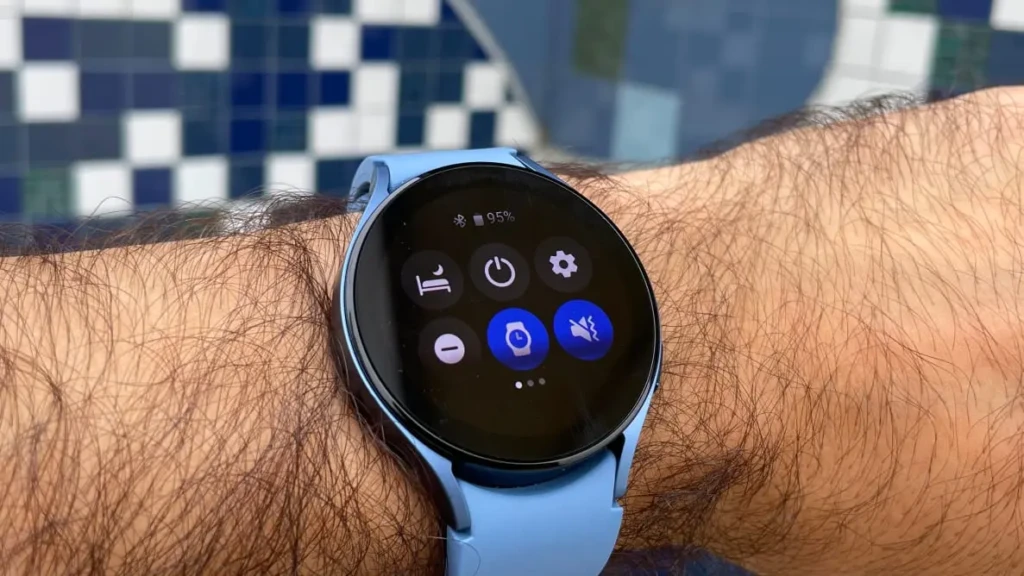 A Samsung Watch 5 in light blue worn on a person’s wrist, displaying the quick settings menu with icons for sleep tracking, power, settings, and Do Not Disturb, set against a blurred blue-and-white tiled background, illustrating real-world usability and intuitive controls.