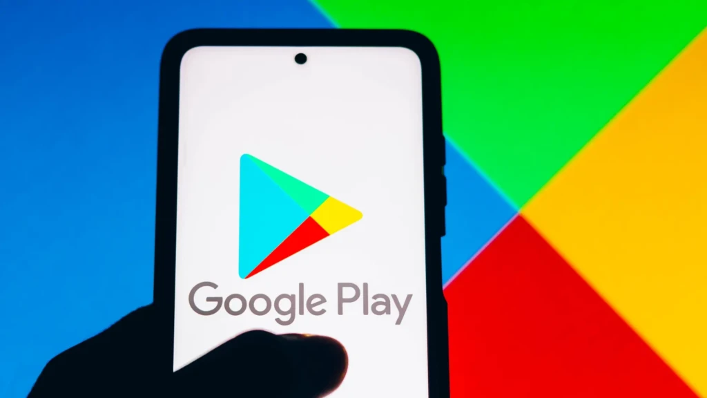 A hand holding a smartphone displaying the Google Play logo on its screen, set against a vibrant multicolored geometric background, emphasizing the brand’s identity and accessibility as the primary gateway to Android apps and digital services.