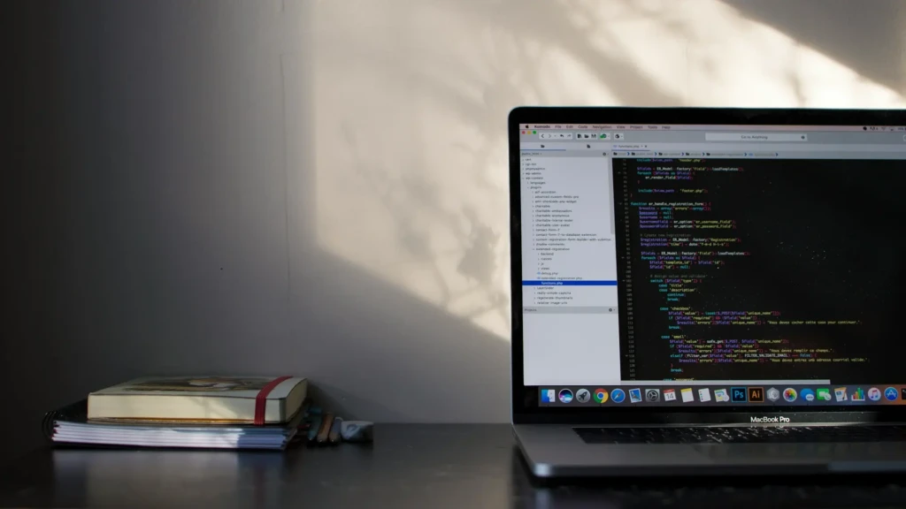 A MacBook Pro on a dark desk showing PHP code in a dark-themed editor, with sunlight casting leaf shadows on the wall beside stacked notebooks and pencils, suggesting a quiet, thoughtful coding session in a personal or home office environment.