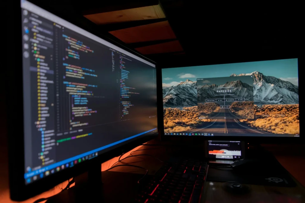 A dual-monitor developer workstation in a dimly lit room, with one screen showing syntax-highlighted code and the other displaying a desktop wallpaper with a “PAPERIZE” app overlay, complemented by a red-backlit keyboard and ambient lighting, evoking a late-night coding session.