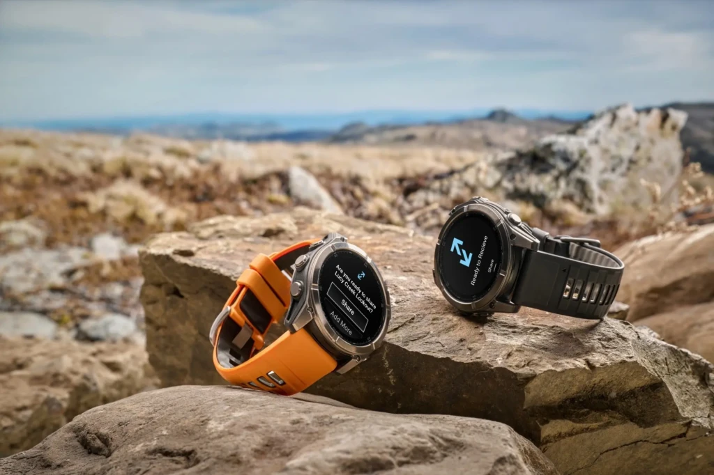Two Garmin Fenix 8 watches, one with an orange strap and one with a gray strap, resting on a rocky mountaintop. Their screens display location-sharing features (“Ready to Receive” and “Are you ready to share Lucy Creek Lookout?”), demonstrating safety and connectivity in remote environments