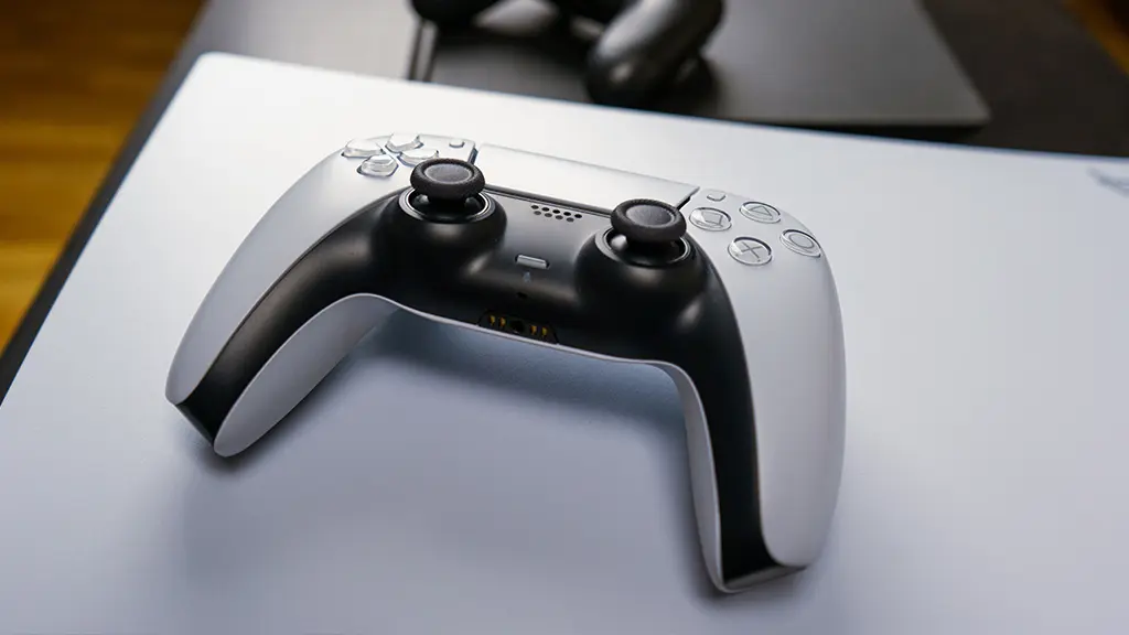 A PlayStation DualSense controller lying flat on a white desk mat, showcasing its sleek black-and-white design, touchpad, speaker grille, and USB-C port, ideal for charging or connecting to devices in a clean, minimalist setup