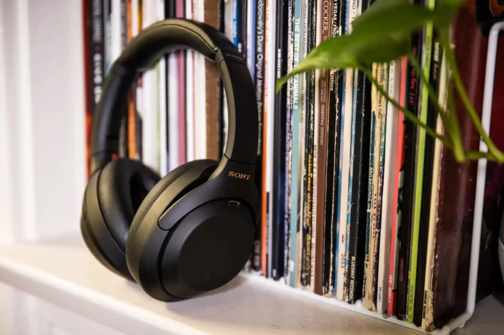A black Sony WH-1000XM4 headset placed on a white shelf beside a collection of vinyl records and a green plant, suggesting a love for high-fidelity audio and blending modern tech with classic music culture in a cozy home environment.