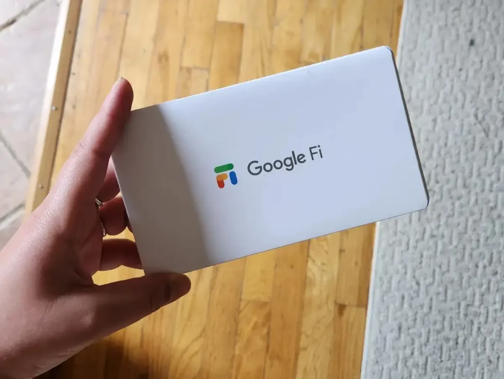 A hand holding a white Google Fi welcome card featuring the colorful “Fi” logo and text “Google Fi,” set against a warm wooden floor and gray rug, highlighting the tangible onboarding experience for new users signing up for the mobile service.