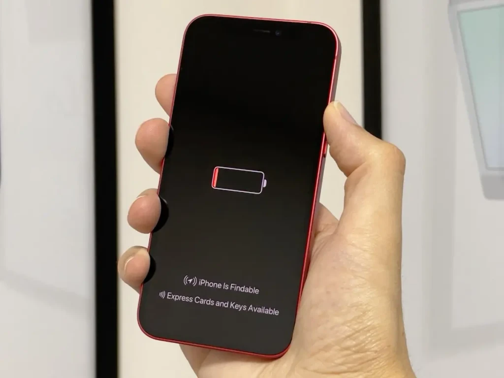 A hand holding a red iPhone with its screen displaying a low battery icon and the message “iPhone Is Findable,” indicating it can still be located even when powered down, highlighting Apple’s advanced tracking feature for lost devices.