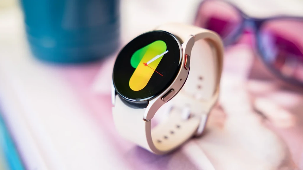 A Samsung Watch 5 in beige strap and rose gold case resting on a soft pink surface beside sunglasses and a water bottle, displaying a colorful gradient analog face, showcasing its fashion-forward appeal and integration into a stylish, active daily routine.