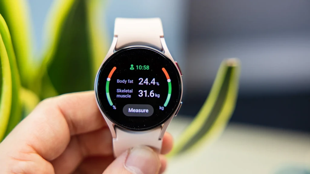 A hand holding a Samsung Galaxy Watch displaying its body composition screen, showing 24.4% body fat and 31.6 kg skeletal muscle with color-coded progress bars, highlighting advanced health tracking features against a blurred green plant background.