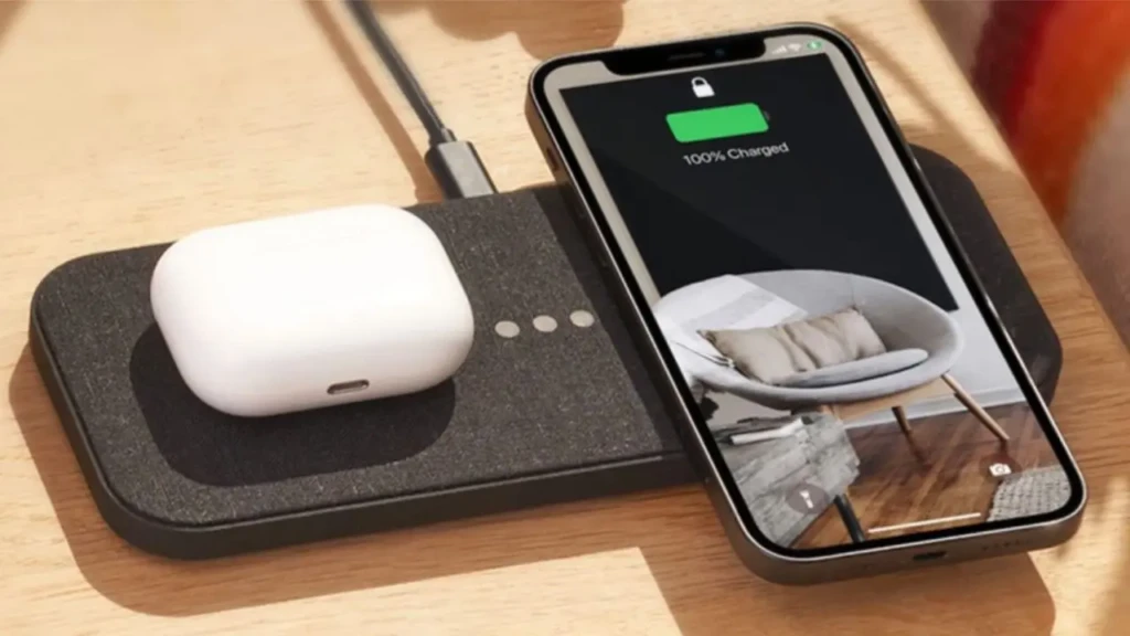 An iPhone displaying “100% Charged” while charging on a black multi-device wireless pad beside a white AirPods case, set on a wooden desk, illustrating seamless, simultaneous charging for Apple devices in a clean, everyday workspace environment.