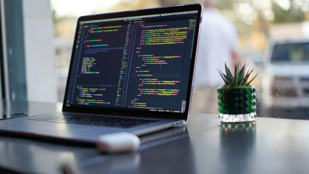 A MacBook Pro open on a reflective black table next to a small green potted succulent, displaying HTML/CSS/JavaScript code in a dark IDE, evoking a clean, minimalist coding environment with natural elements and soft background lighting.