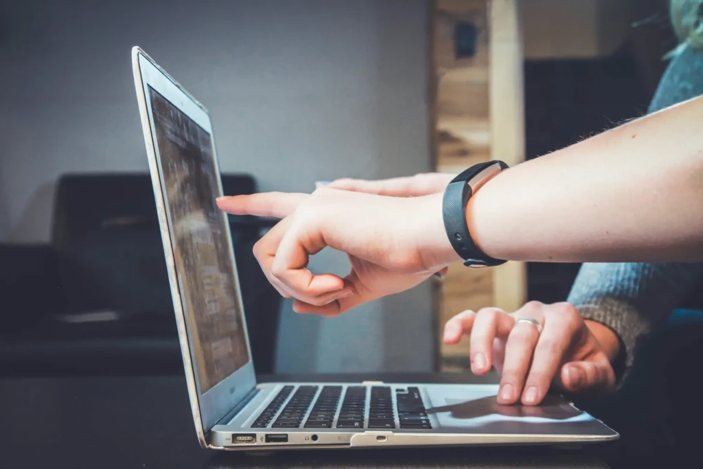 A close-up of hands interacting with a MacBook, one finger tapping the screen while the other rests on the trackpad, with a fitness tracker on the wrist, showcasing intuitive multitasking and seamless user experience while working on a laptop.