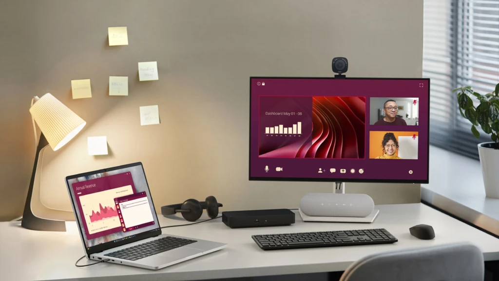 A modern home office setup featuring an HP laptop connected to an external monitor displaying a video conference with two participants and a shared dashboard. The desk includes a webcam, speaker, headphones, keyboard, mouse, and a warm desk lamp with sticky notes on the wall behind