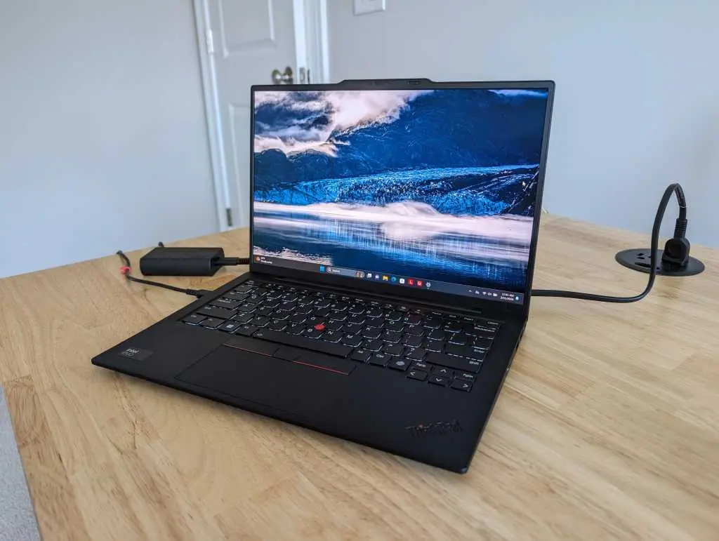 A black Lenovo ThinkPad laptop open on a light wood desk, displaying a scenic mountain and lake wallpaper. The laptop is connected to a power adapter, with a cable running to a wall outlet in the background