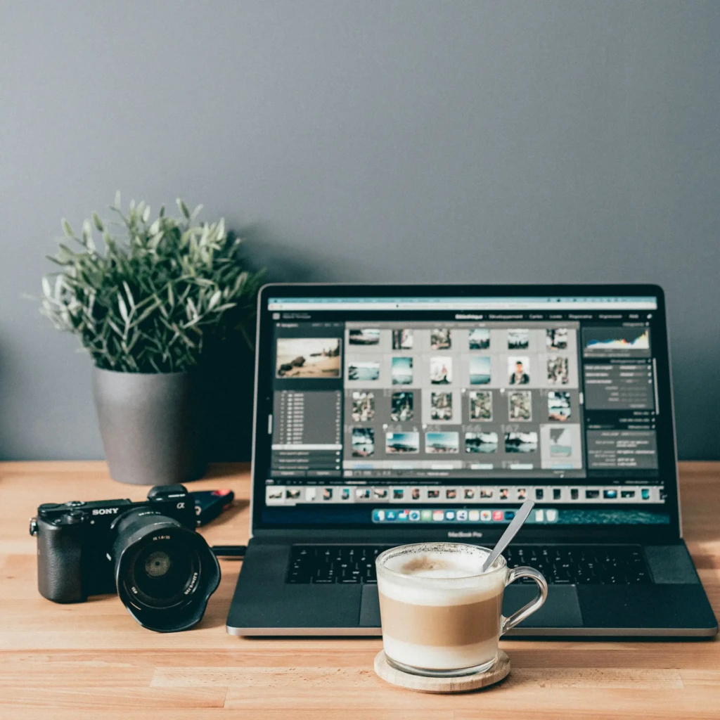 A cozy workspace scene with a MacBook Pro running photo editing software displaying a grid of thumbnails, beside a Sony camera and a glass of coffee on a wooden desk, illustrating a professional or enthusiast photographer’s editing setup in a calm, productive environment.