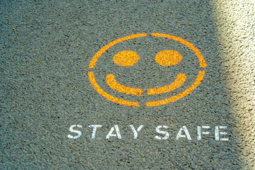 An orange smiley face painted on asphalt pavement with the words “STAY SAFE” stenciled below in white, conveying a cheerful, community-focused safety reminder in public spaces, often used during health or social awareness campaigns.