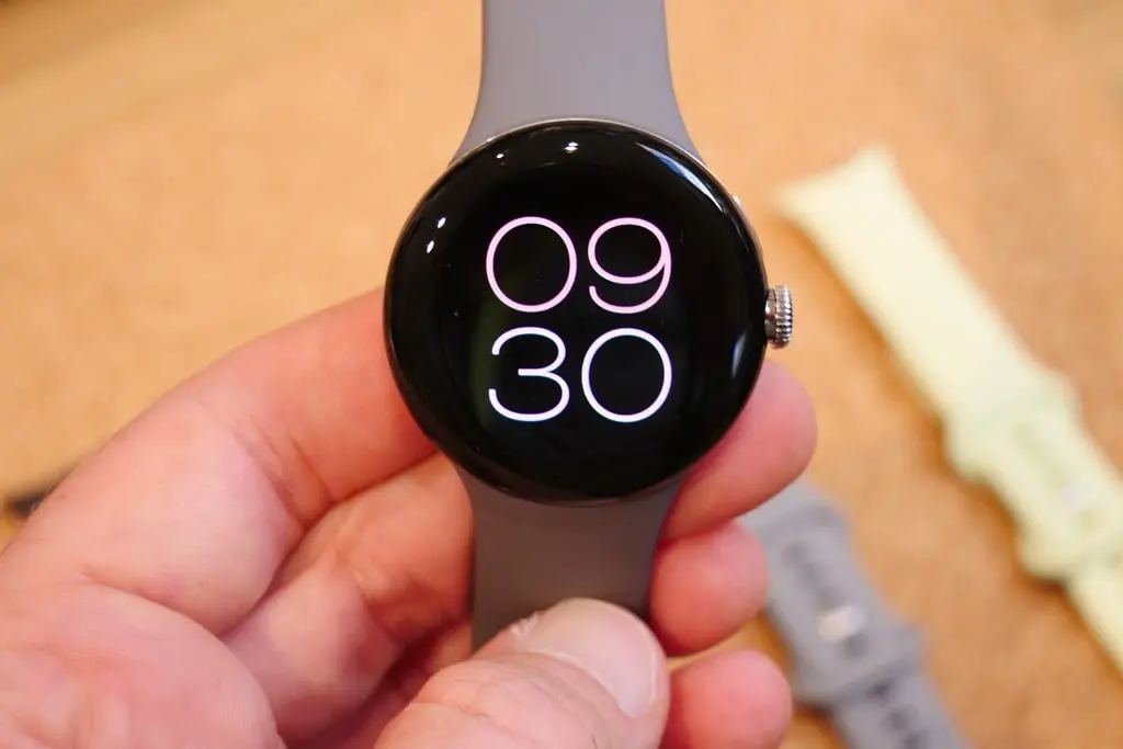 A hand holding a Google Pixel Watch with a gray silicone strap, displaying a minimalist digital watch face showing “09:30” in glowing white numerals against a black background, with spare watch bands blurred in the background, highlighting its sleek design and customizable accessories.