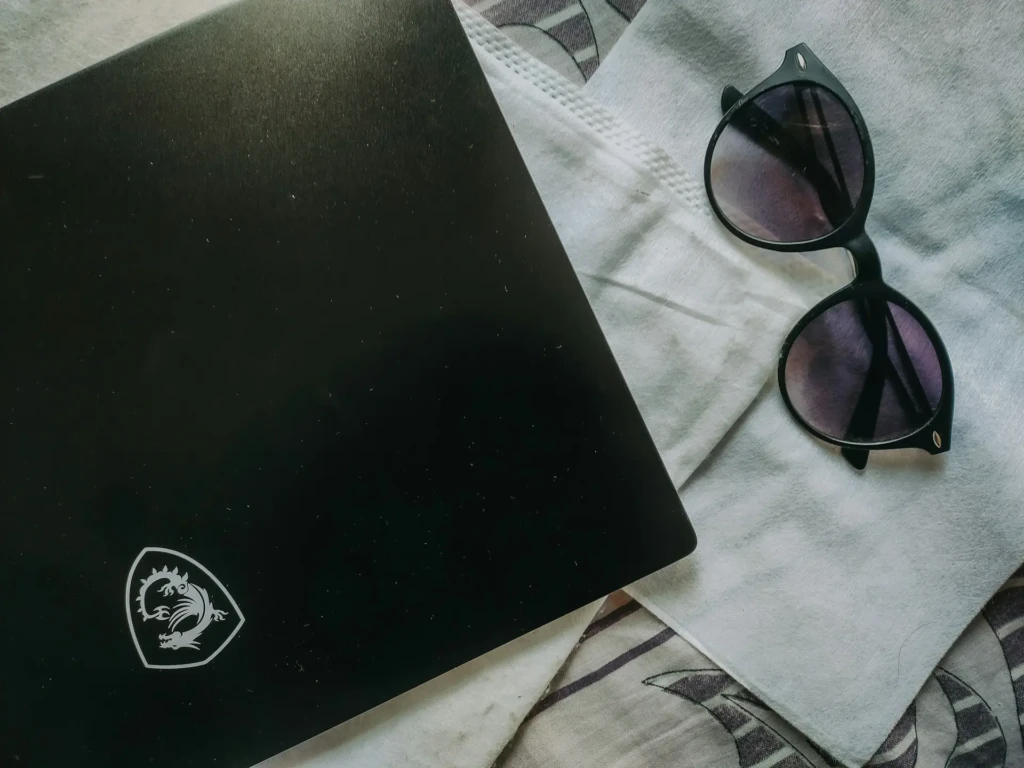 A sleek black laptop with a dragon logo sits beside stylish black sunglasses on a textured white cloth and patterned background.