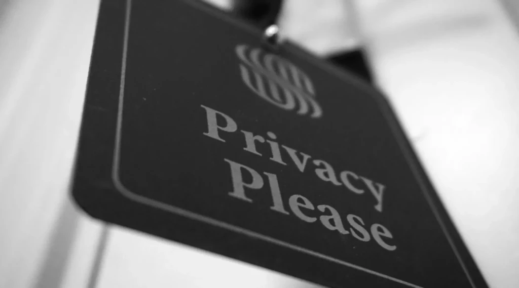 A close-up black-and-white photo of a door hanger with a dollar-sign-like symbol and the words “Privacy Please” printed in bold serif font, symbolizing user privacy, data protection, or confidentiality in digital contexts.