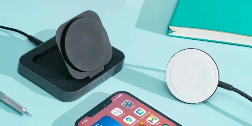 A Zens wireless charging station with a black magnetic puck and base displayed next to a white circular Qi charger, both arranged on a light blue surface beside an iPhone and notebook, showcasing versatile charging options for iPhone users in a modern, minimalist setup.