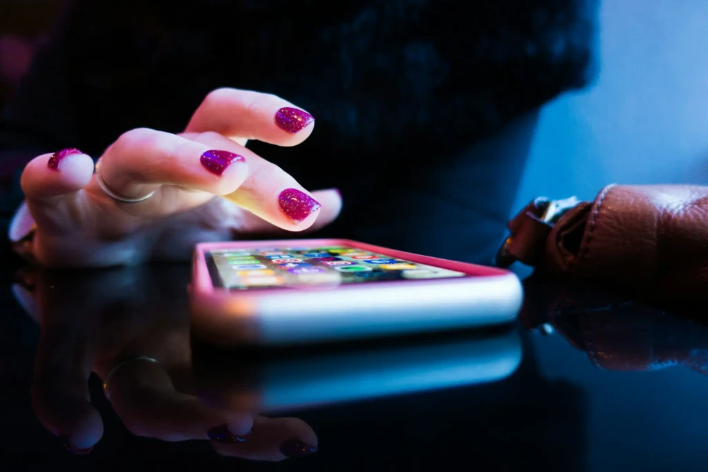 A close-up of a hand with glittery purple nails tapping a smartphone on a glossy surface, with a brown wallet partially in view.