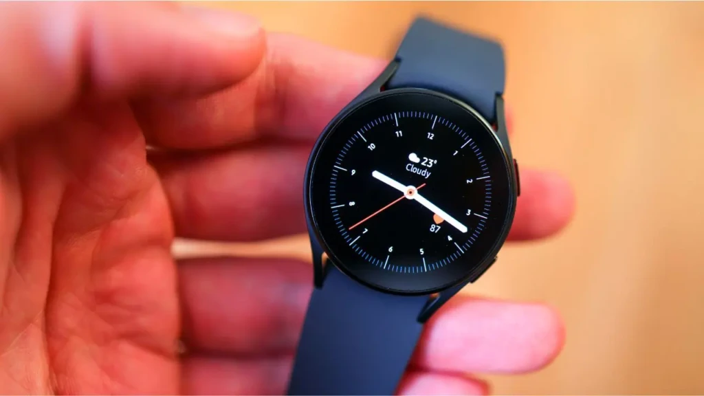 A hand holding a Samsung Watch 5 with a black silicone strap and black bezel, showing an analog watch face with weather (23°C, cloudy) and heart rate (87 bpm), highlighting its health monitoring features and classic design in a warm, lifestyle-oriented setting.