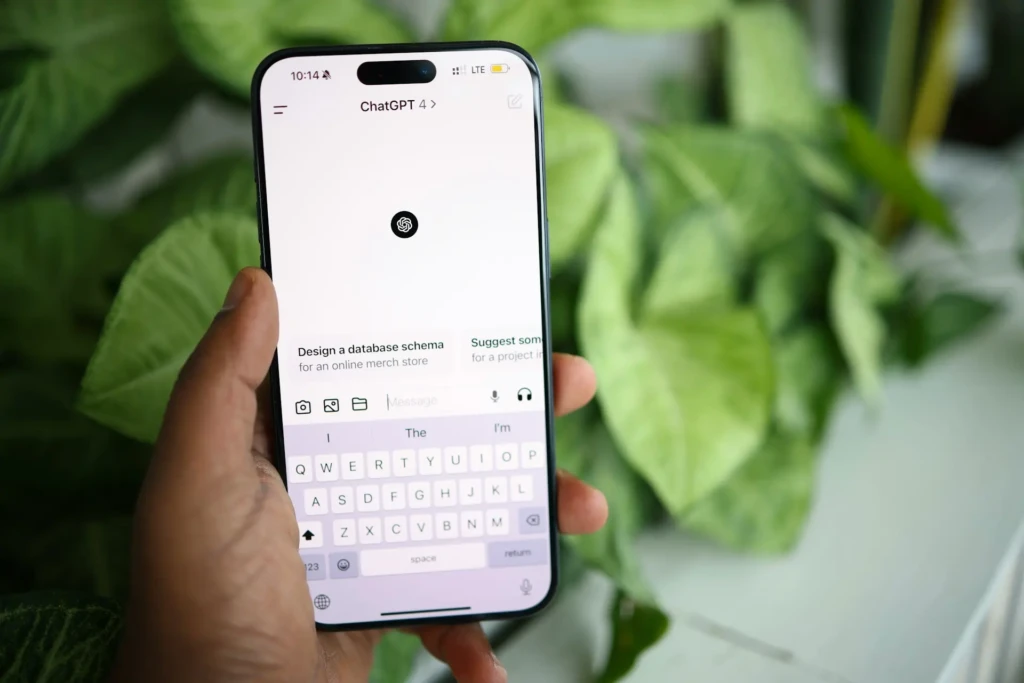 A hand holding an iPhone displaying the ChatGPT 4 app interface with suggested prompts like “Design a database schema for an online merch store,” set against a blurred green leafy background, showcasing real-world usage and conversational interaction in a natural, everyday setting.
