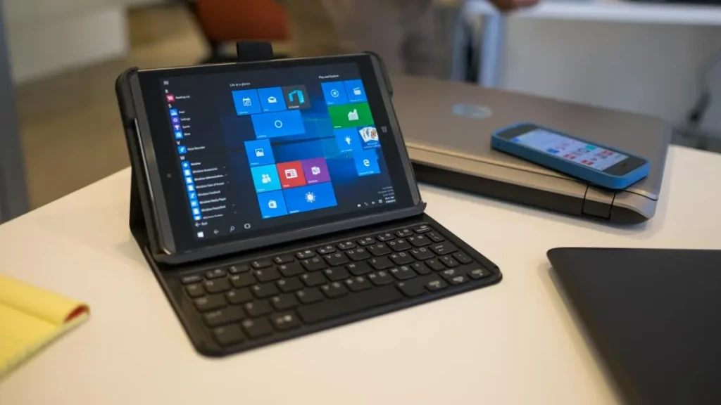 A black HP tablet with a detachable keyboard dock, displaying the Windows Start menu on its screen. It sits on a white desk next to a closed laptop and a blue smartphone, suggesting a mobile productivity setup