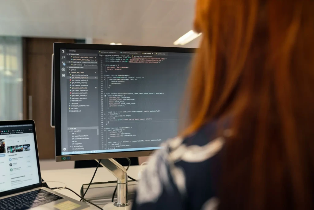 A developer with long red hair reviewing JavaScript code on a large Samsung monitor, with file explorer and function outlines visible, seated at a desk with a laptop open to a Twitter feed, suggesting collaborative or API-focused development work in a modern office.