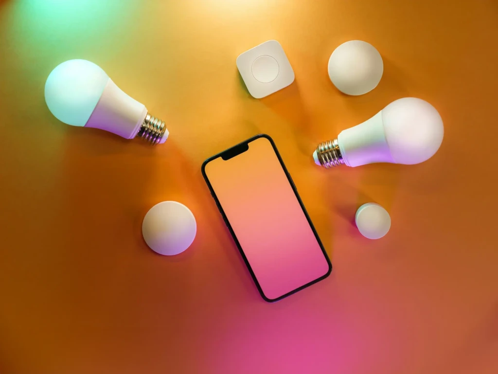 A top-down view of multiple smart bulbs and a smartphone arranged on a warm orange surface, with colorful ambient light cast by the bulbs, showcasing compatibility, variety, and vibrant color-changing capabilities for modern smart home setups.