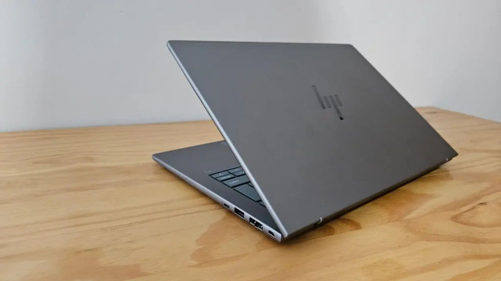 A sleek gray HP laptop partially open on a light wood table, viewed from the rear-left angle. The HP logo is subtly etched on the lid, and multiple ports are visible along the side, emphasizing connectivity and minimalist design
