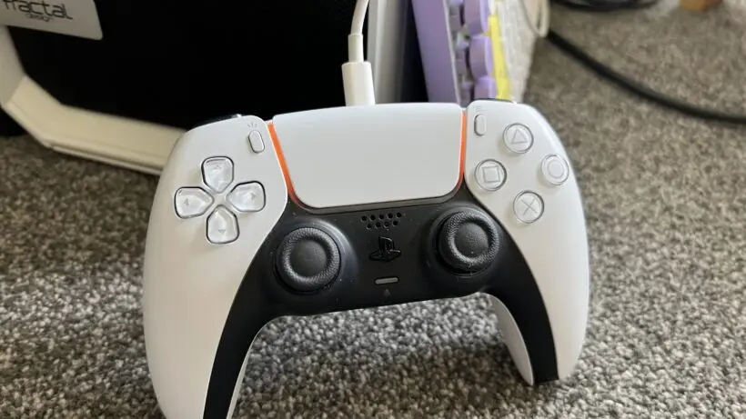 A white PlayStation DualSense controller resting on a carpeted surface, connected via USB-C cable. The background includes a Fractal Design case and keyboard, indicating a PC gaming setup or charging scenario