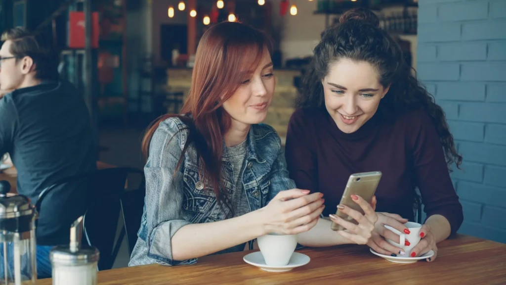 Two women smiling while looking at a smartphone together in a cozy café, holding coffee cups, suggesting collaborative or social use of mobile apps, possibly including security tools like Google Authenticator, in everyday casual settings.