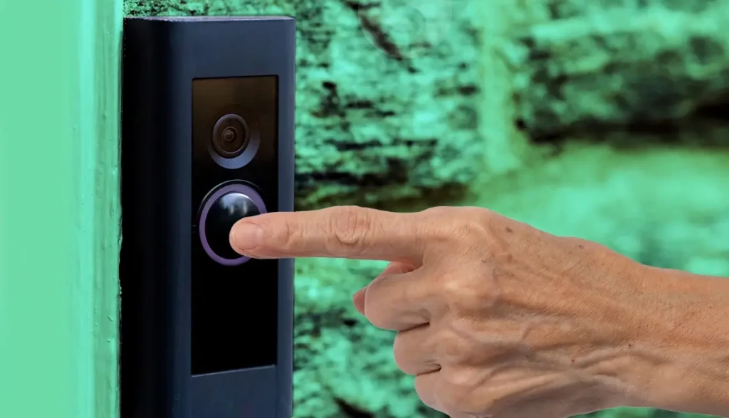 A close-up of a finger pressing the illuminated button on a black smart video doorbell mounted on a textured green wall, showcasing its sleek design, responsive touch interface, and intuitive user interaction for welcoming or alerting residents.
