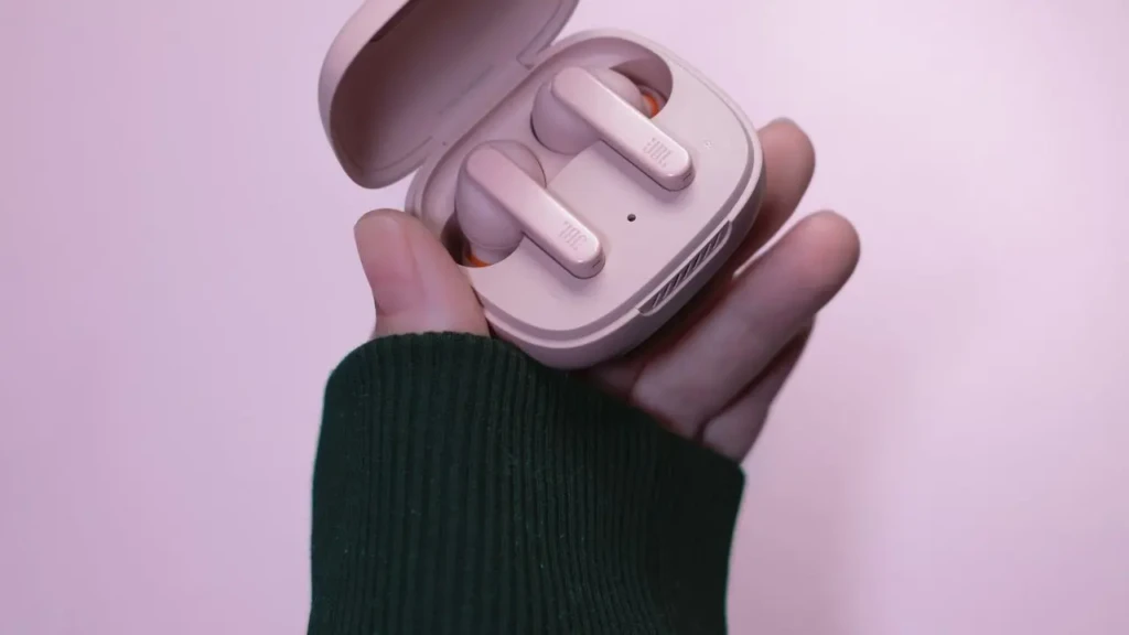 A hand holding a light pink JBL true wireless earbud case open to reveal two matching earbuds, set against a soft lavender background, showcasing its compact, fashion-forward design and portability for everyday use.