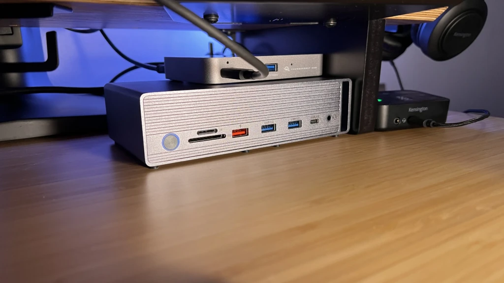A silver textured docking station with front-facing ports (TF/SD card slots, red USB-C charging port, USB 3.2, headphone jack) placed under a desk next to a Thunderbolt hub and a Kensington audio adapter, with blue ambient lighting, emphasizing organized cable management and multi-hub integration in a compact desktop setup.