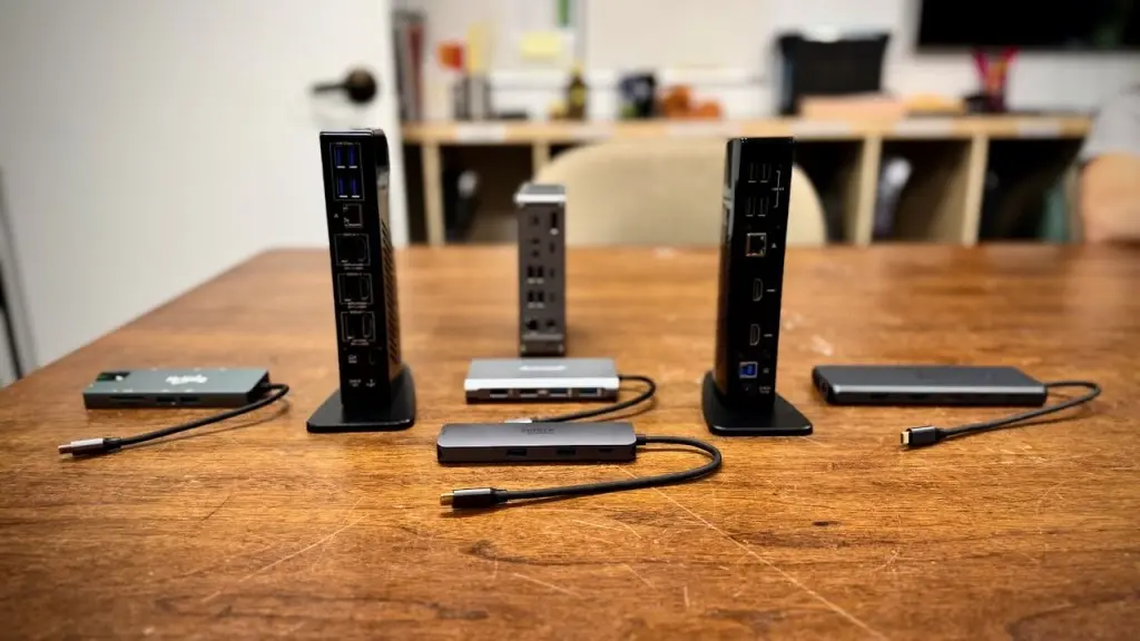 Five different docking stations arranged on a wooden table, ranging from compact horizontal hubs to tall vertical docks, each with visible cables and ports, illustrating a curated selection for workspace expansion and peripheral integration.