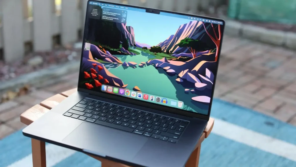 A space-black MacBook Pro open on a wooden stool outdoors, displaying a vibrant animated landscape wallpaper with app icons in the dock and a notification panel visible, highlighting its vivid display and portability in a natural, sunlit setting.