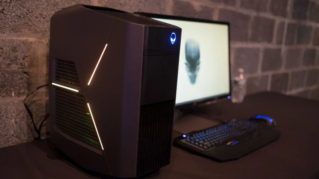 A classic Alienware Aurora desktop with angular design, green-and-orange LED strips, and the glowing blue alien logo, positioned next to a monitor showing the Alienware logo wallpaper, a backlit keyboard, and mouse on a dark desk against a brick wall, evoking a dedicated, high-end gaming setup.