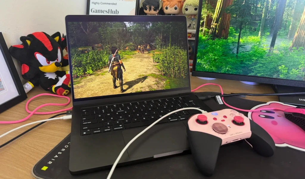 A laptop displays a gaming scene with a character walking on a path, alongside a pink game controller and a plush toy.