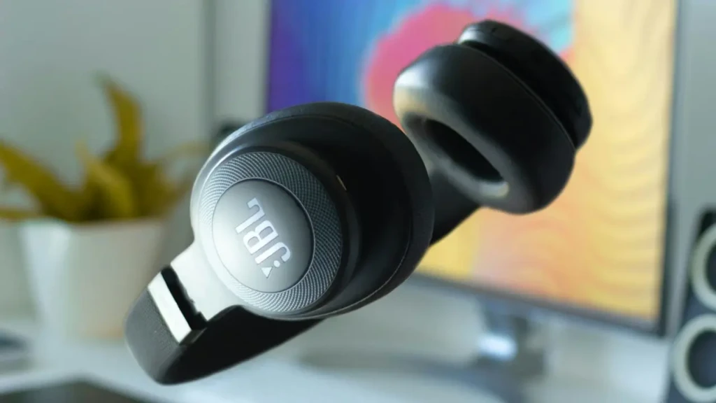 A black JBL over-ear headphone suspended mid-air in front of a colorful monitor, with soft-focus plants and decor in the background, highlighting its sleek design, textured earcup finish, and integration into a vibrant, creative home-office environment.